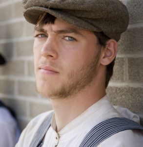 Young man in 20s garb leans against a wall