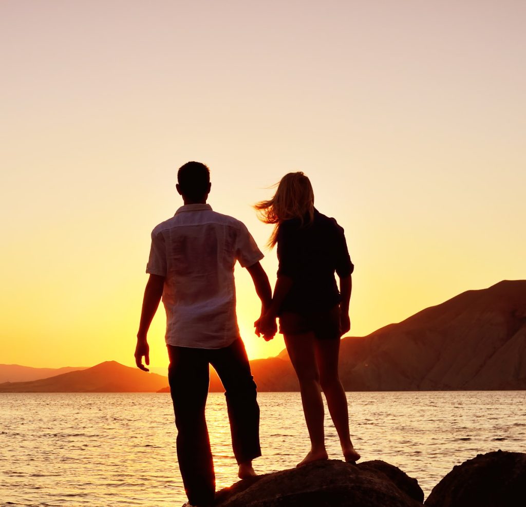 A couple stands at the sea at sunset, hand in hand.